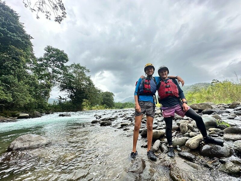 Here is a picture of two people standing in a river. They are both wearing life vests and helmets, suggesting they are about to go rafting or have just finished. The background shows a cloudy sky and lush greenery, indicating a natural environment.